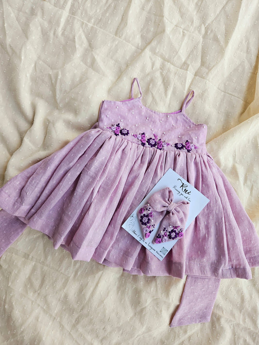 Flatlay of Lovely Lavender dress with matching embroidered bow on cream fabric.