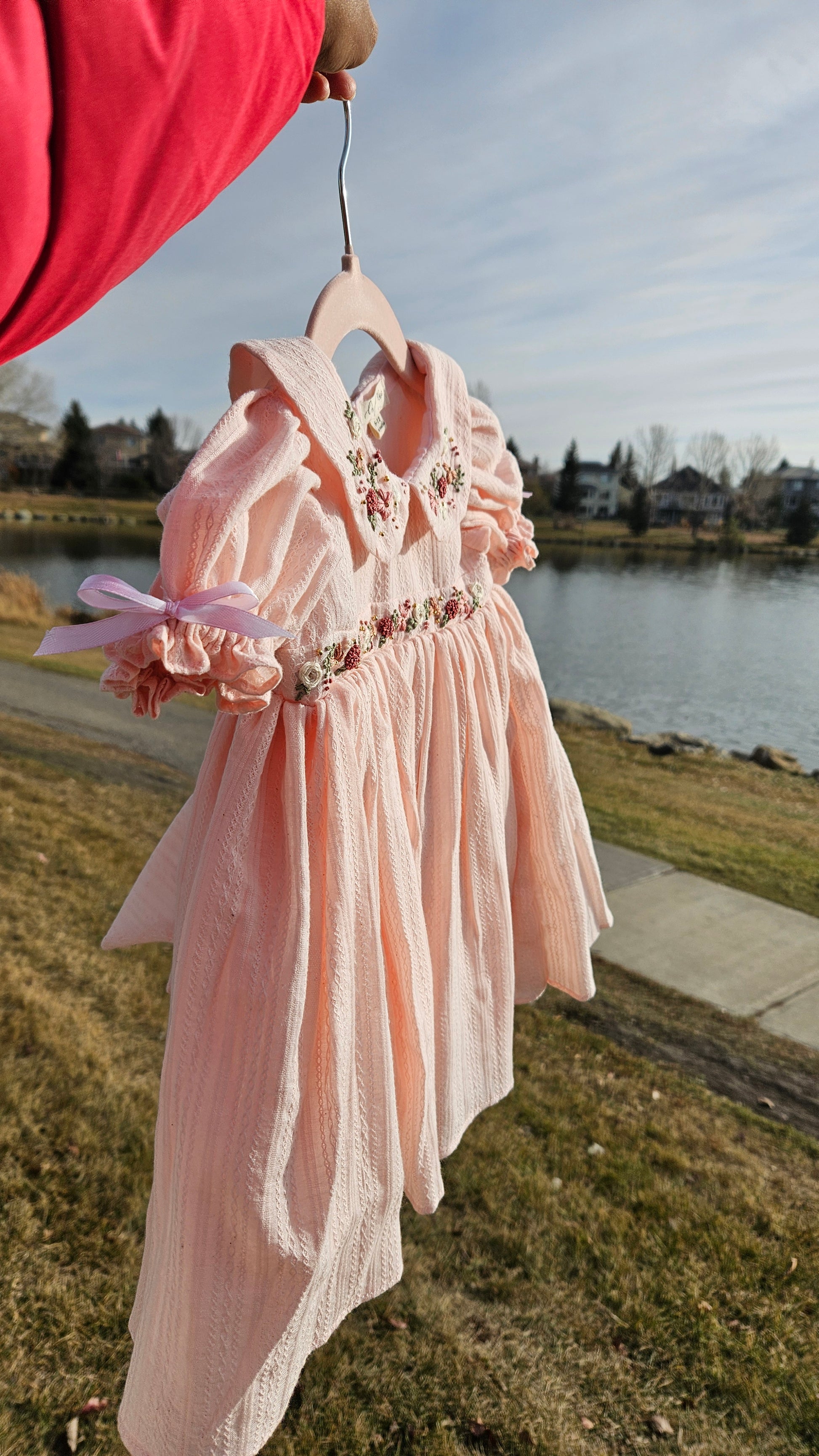 Peachy Peep embroidered baby dress held outdoors near a lake.