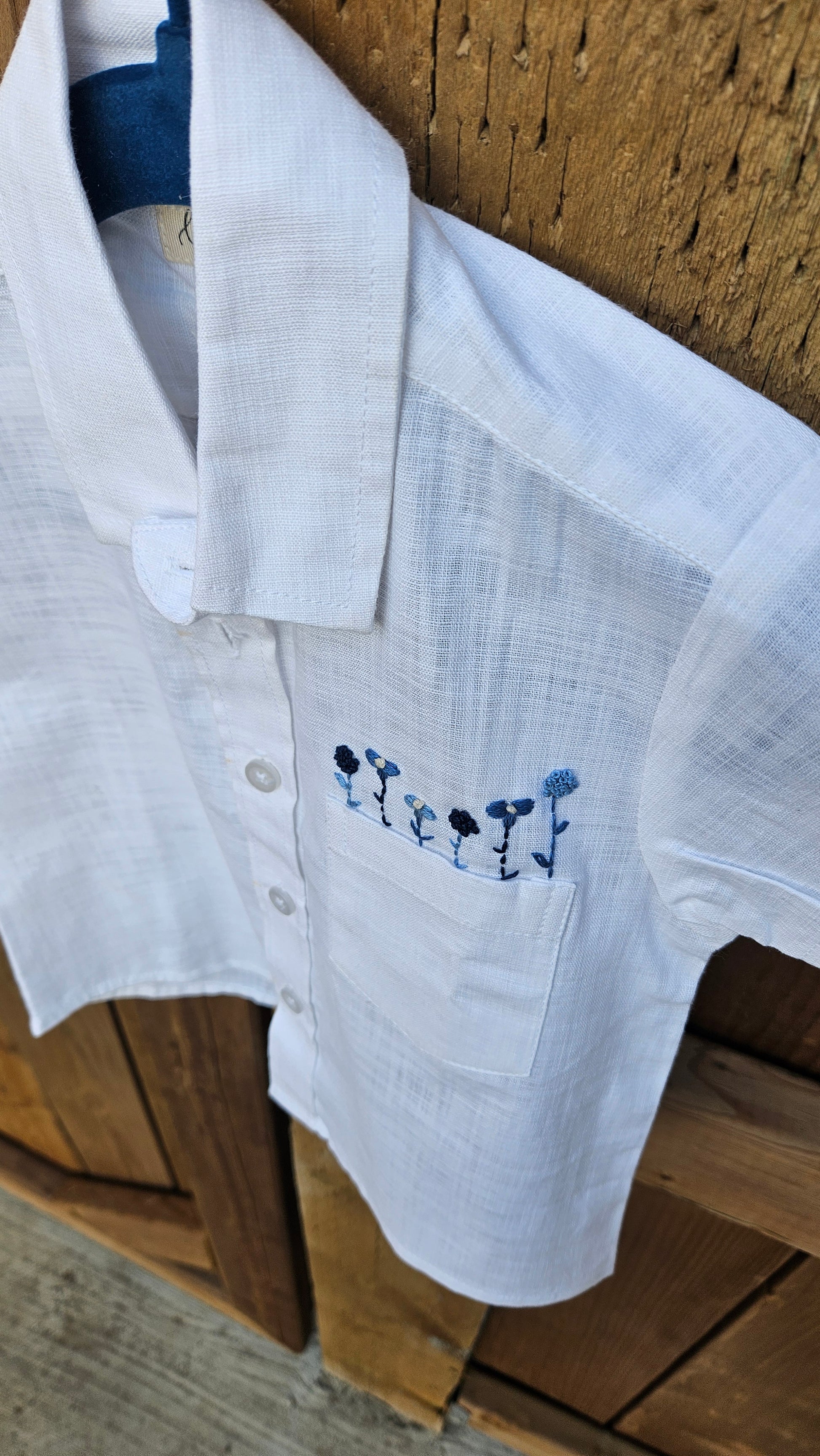 White cotton-linen boys’ shirt with blue embroidered flowers hung on a wooden fence.