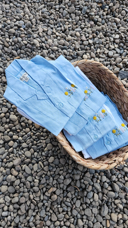 Sky-blue embroidered boys’ shirts placed in a basket on pebbles.
