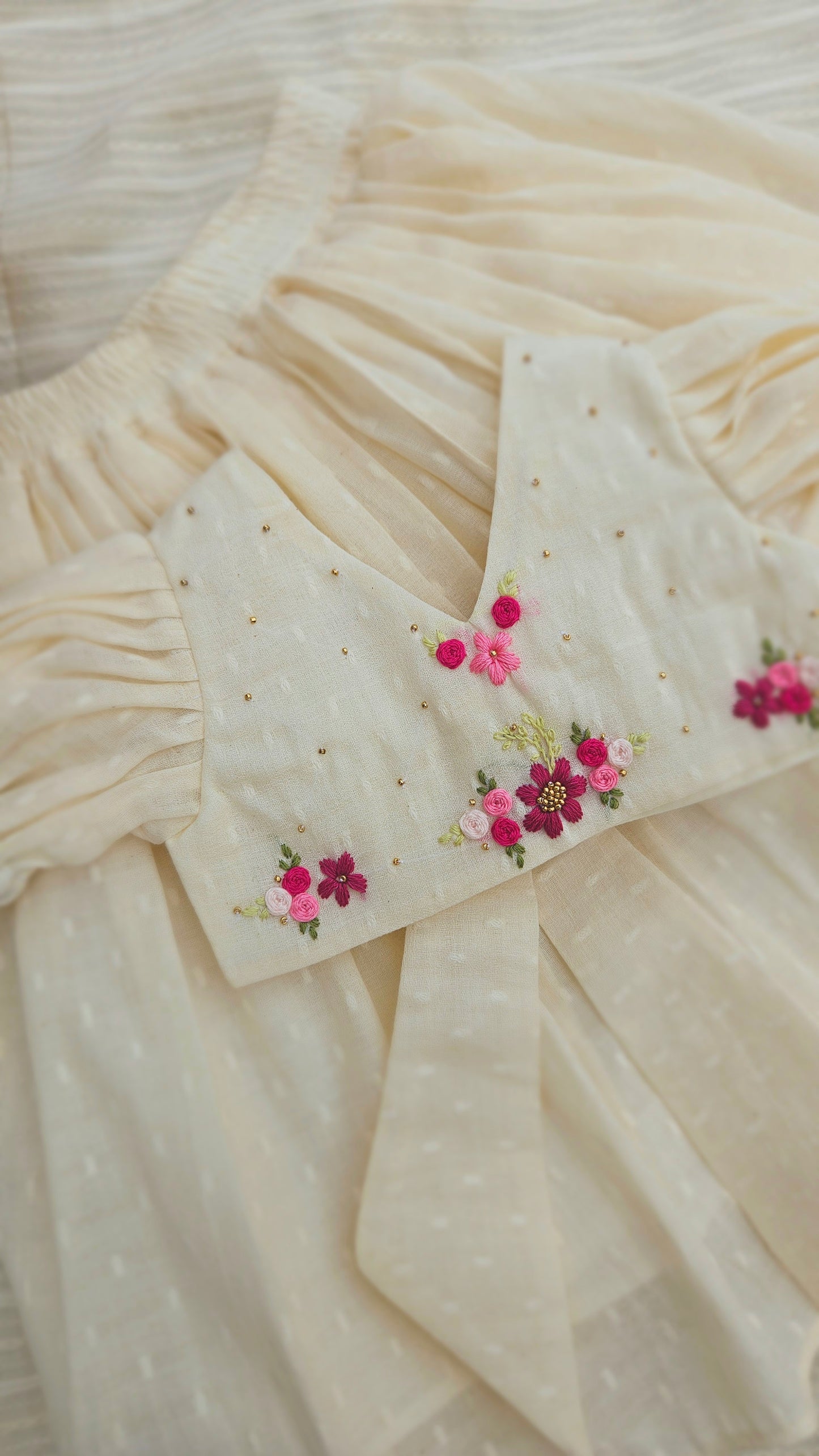 Cream onam-inspired kids lehenga set with hand-embroidered pink and magenta floral motifs and gold bead detailing, displayed on a soft cream fabric background.