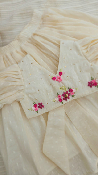 Cream onam-inspired kids lehenga set with hand-embroidered pink and magenta floral motifs and gold bead detailing, displayed on a soft cream fabric background.