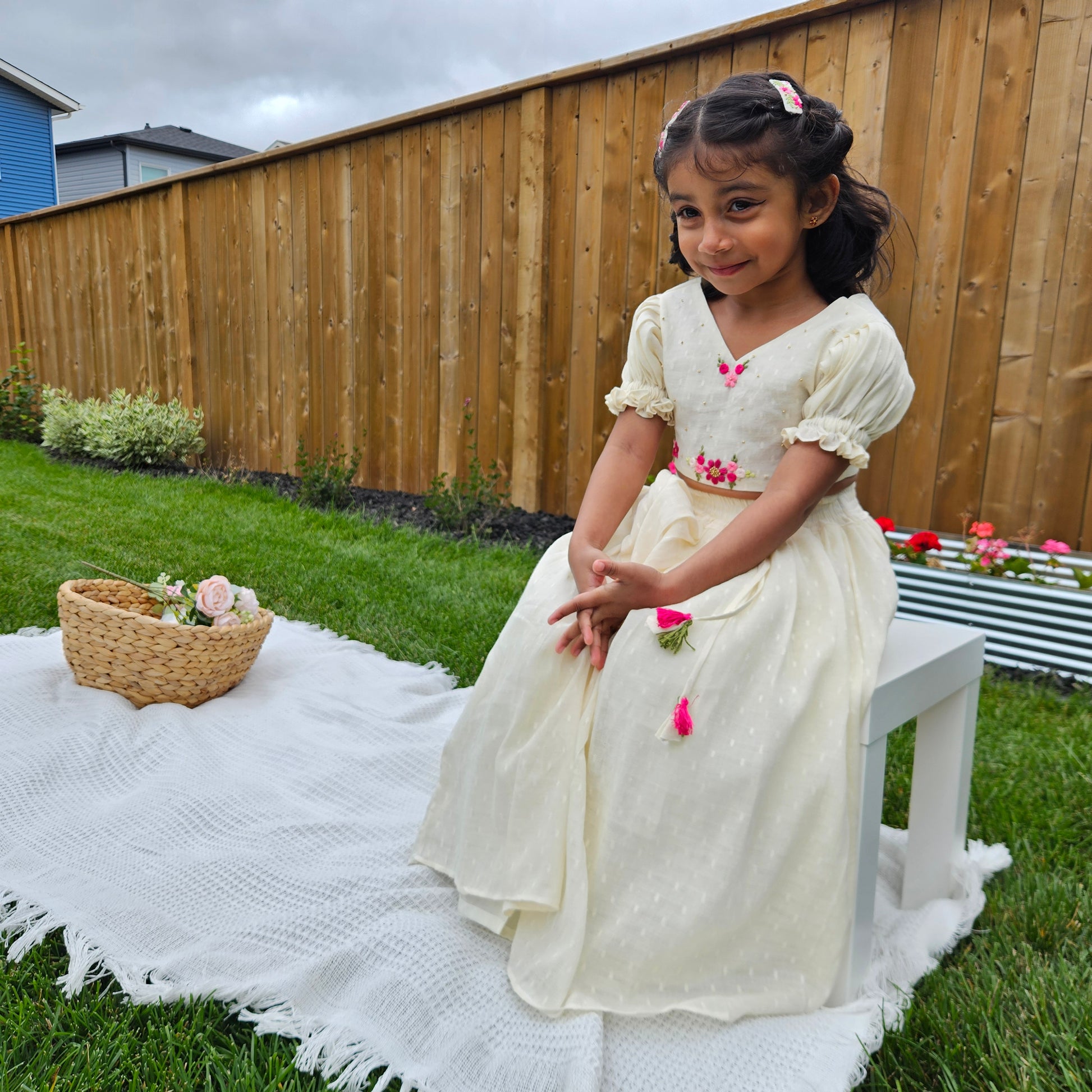 Little girl wearing a cream embroidered lehenga set with bright pink floral work, sitting on a white mat in a garden with a wooden fence behind her.
