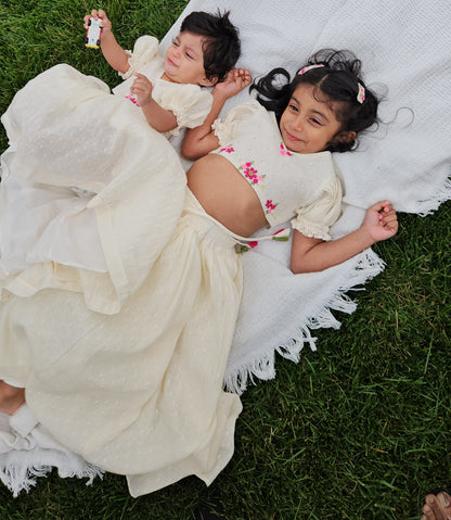 Two little girls wearing matching cream embroidered lehenga sets lying on a white textured blanket on green grass, older sister smiling while younger baby lies beside her.