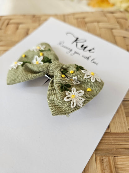 Green floral hair clip on a white card with 'Rae' printed on it, against a woven background.