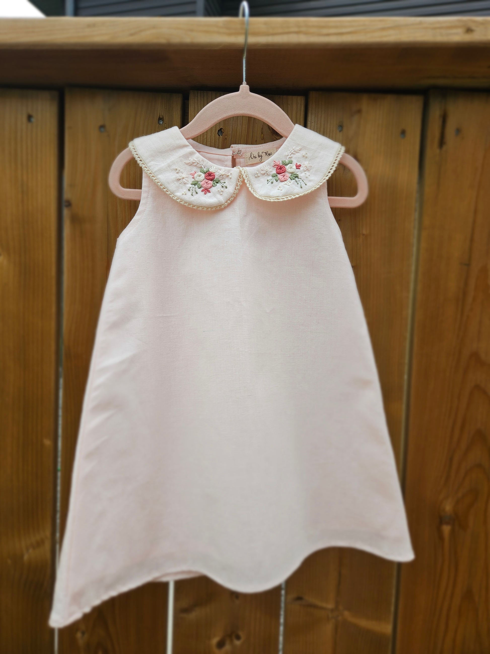 A baby pink cotton linen A-line dress for girls featuring a hand-embroidered Peter Pan collar with delicate pink floral embroidery and scalloped trim. The sleeveless dress has a classic silhouette, making it perfect for birthdays, festive occasions, family outings, and special celebrations.