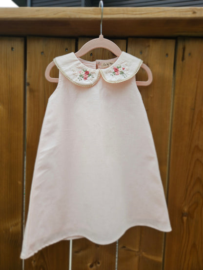 A baby pink cotton linen A-line dress for girls featuring a hand-embroidered Peter Pan collar with delicate pink floral embroidery and scalloped trim. The sleeveless dress has a classic silhouette, making it perfect for birthdays, festive occasions, family outings, and special celebrations.