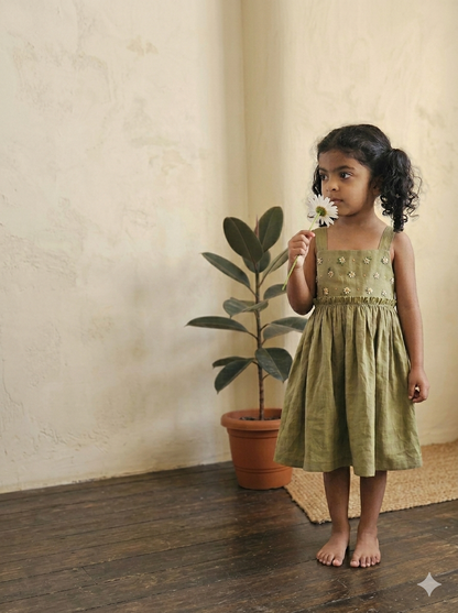 Young girl in a green dress holding a flower next to a potted plant against a beige wall.