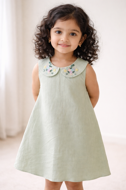 Young girl wearing a light green dress with an embroidered collar against a neutral background